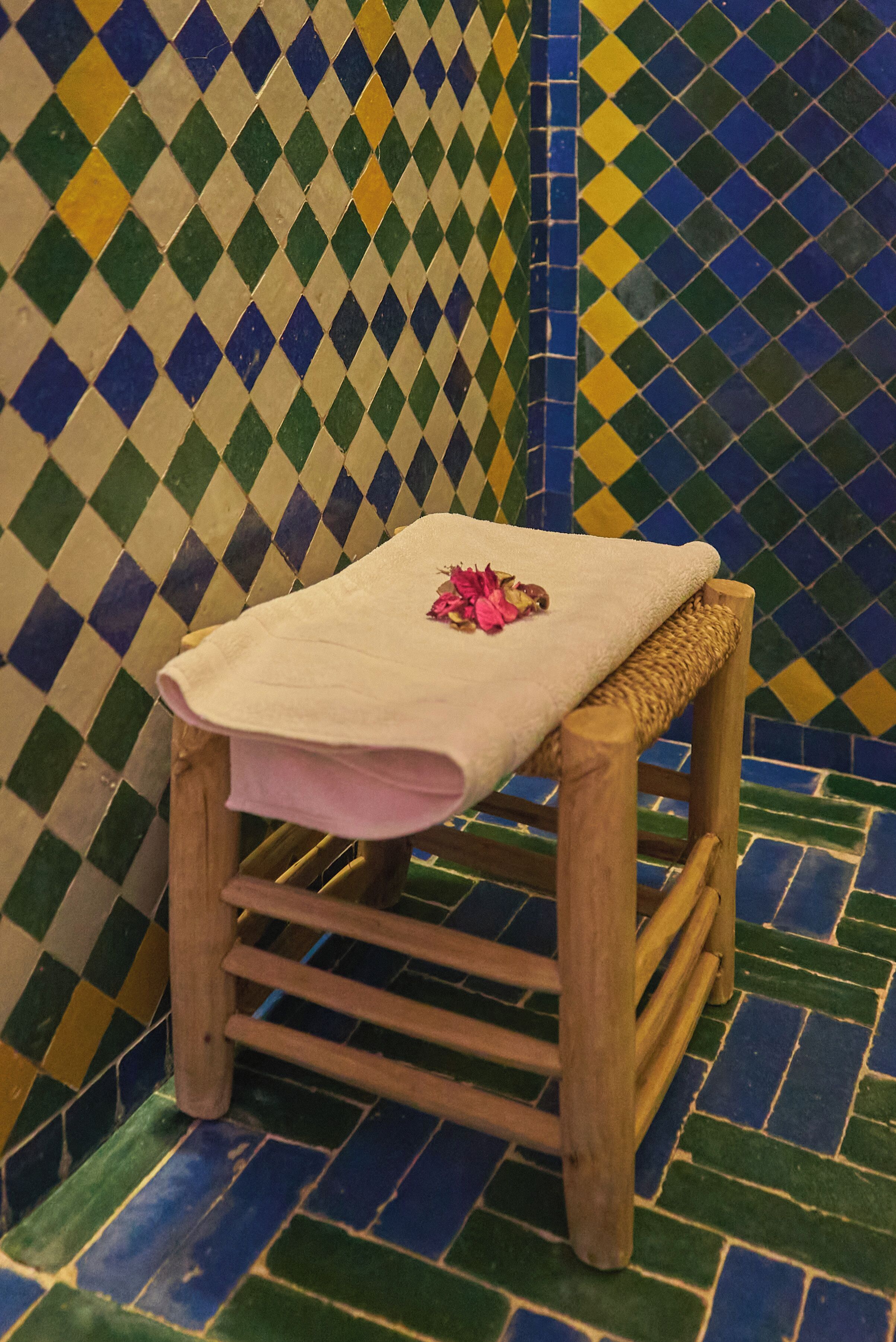 Un taburete de madera con una toalla blanca y flores rosadas encima, apoyado contra una pared de azulejos con diseño a cuadros.