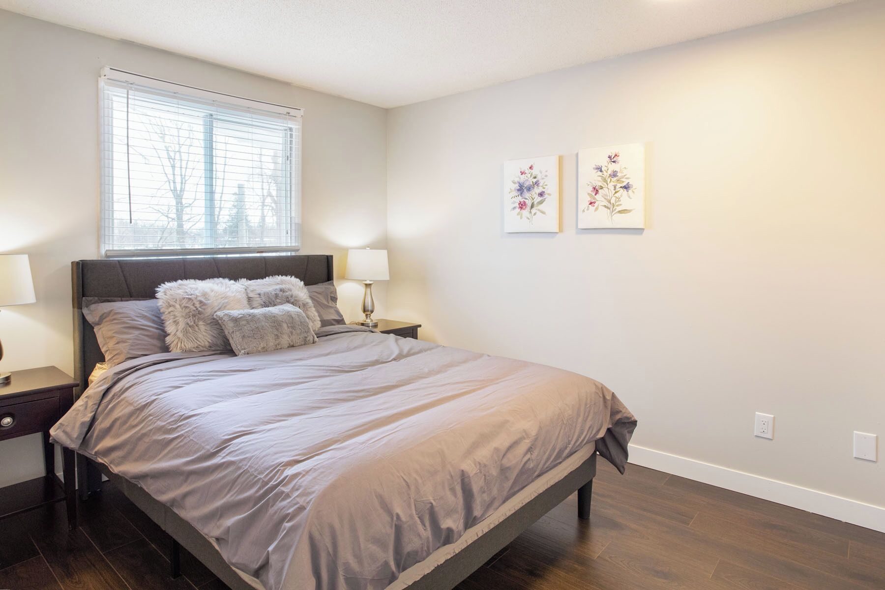 A bedroom with a large bed, two nightstands, a window with blinds, and two floral wall art pieces.