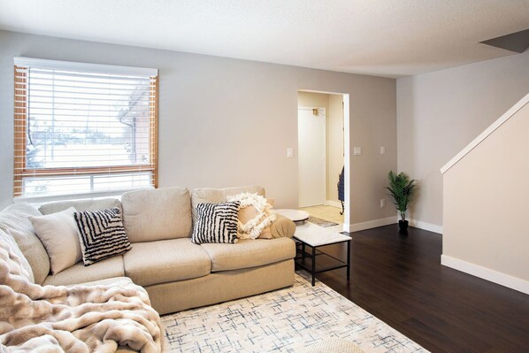 A living room with a beige sofa, patterned pillows, a throw blanket, a small table, and a potted plant.