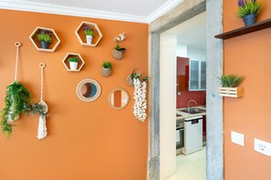 Une pièce avec un mur orange, des étagères hexagonales en bois ornées de plantes, une porte blanche et une cuisine visible à travers une porte ouverte.