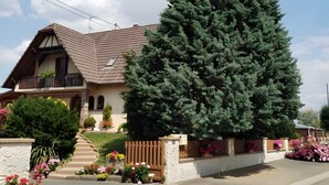 Une maison d’habitation avec un toit en pente, un balcon en bois et un grand arbre à feuilles persistantes dans le jardin.