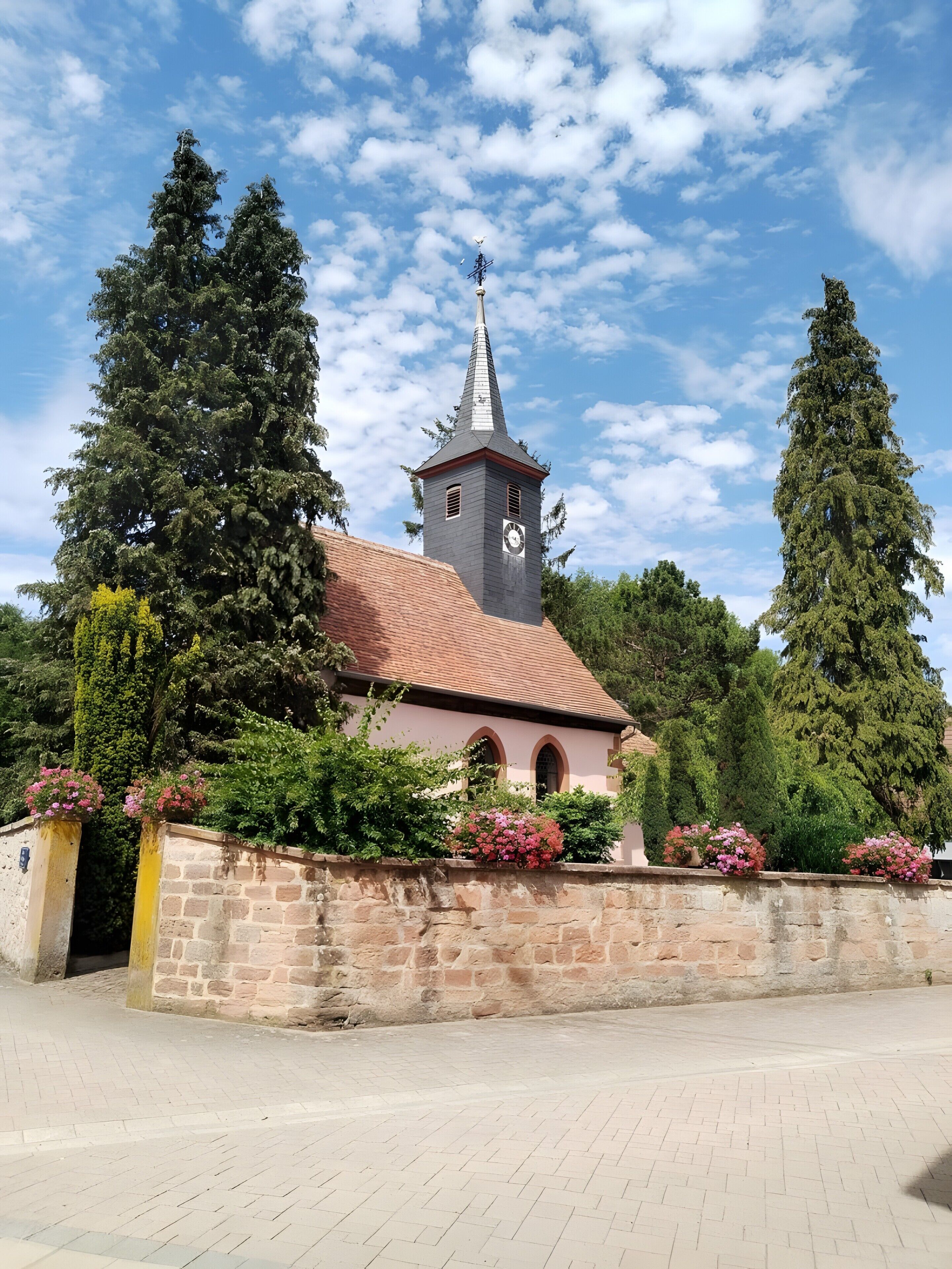 Une église dotée d’un clocher, entourée de murs de pierre et de grands arbres.