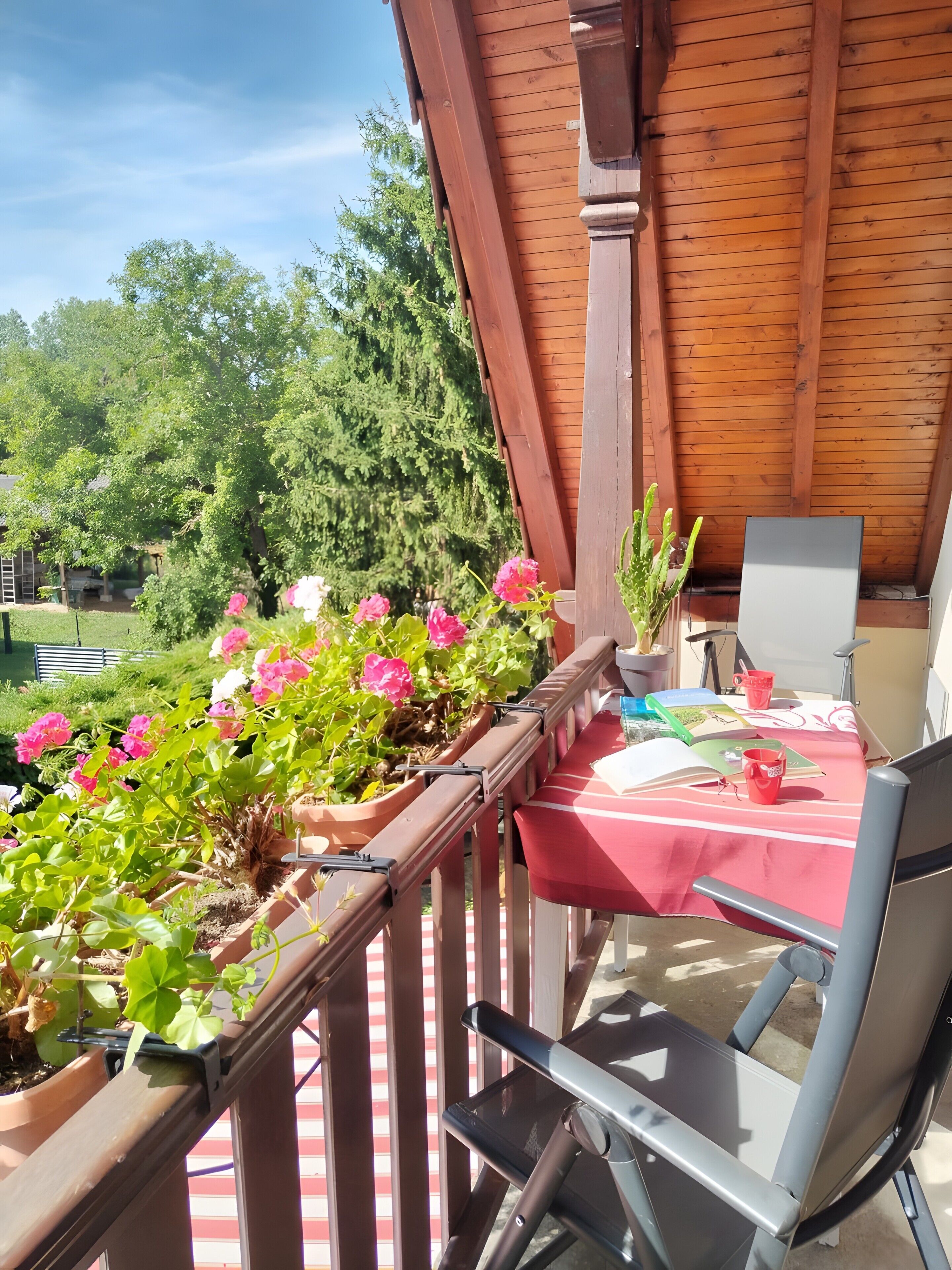 Ein Balkon mit Holzgeländer, einem Tisch mit rotem Tischläufer und Kübelpflanzen mit rosa Blüten.
