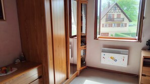 Une pièce avec une armoire en bois, un radiateur, une fenêtre donnant sur une maison et une petite table sur laquelle se trouvent divers objets.