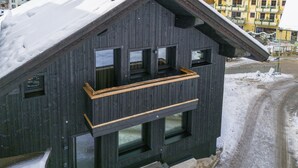 Eine dunkle Holzhütte mit einem Balkon, umgeben von schneebedeckten Bergen und einem Skigebiet im Hintergrund.