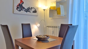A dining area with a wooden table, four chairs, a bowl of fruit, a painting, a lamp, and a window with curtains. - Casa Elena Cozy Stay Steps From Metro Station (Milan)
