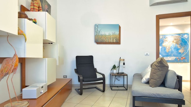 A living room with a sofa, a chair, a small table with a lamp, and a shelf with decorative items. - Casa Elena Cozy Stay Steps From Metro Station (Milan)