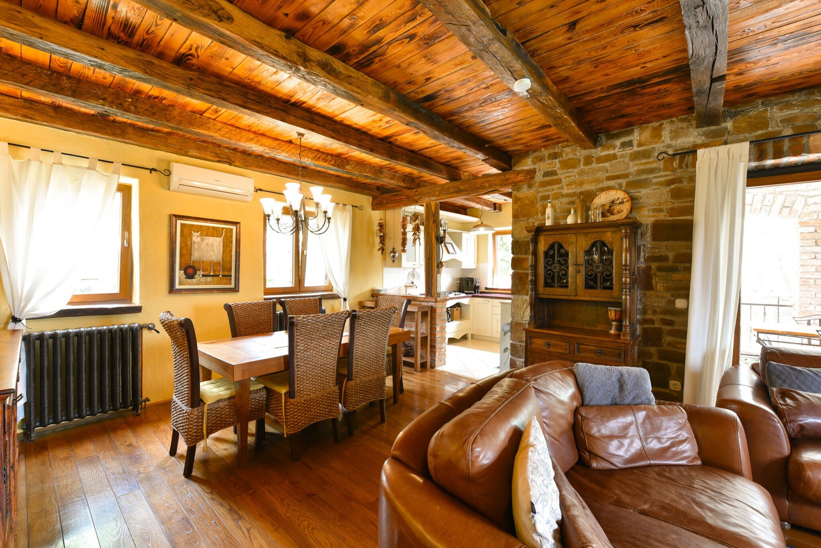 Un intérieur rustique avec un plafond en bois, un coin repas avec des chaises en osier, un canapé en cuir et une cheminée en pierre.