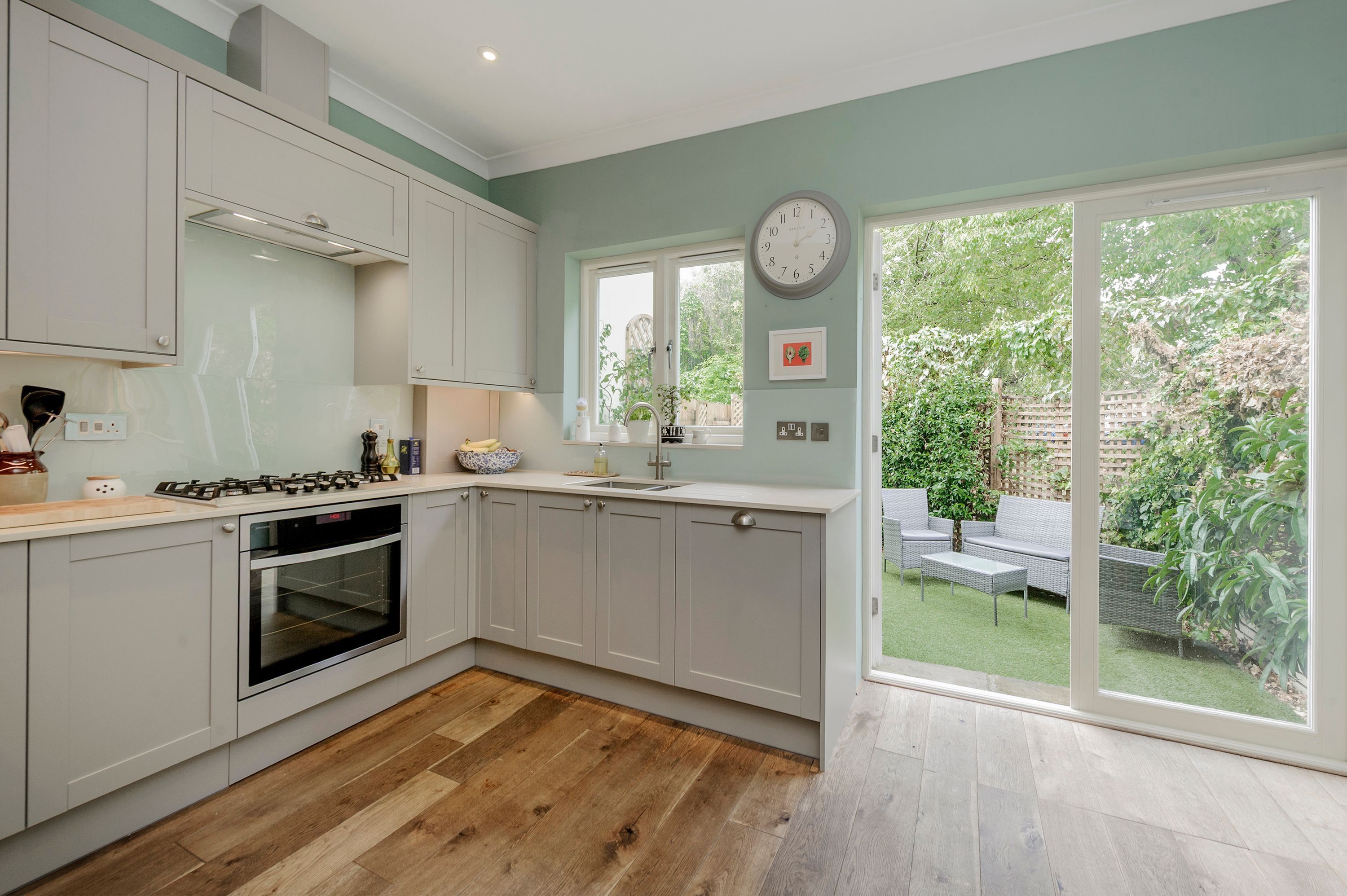 A modern kitchen with white cabinets, a built-in oven, and a large window overlooking a garden. - Wild Roses in London (London)