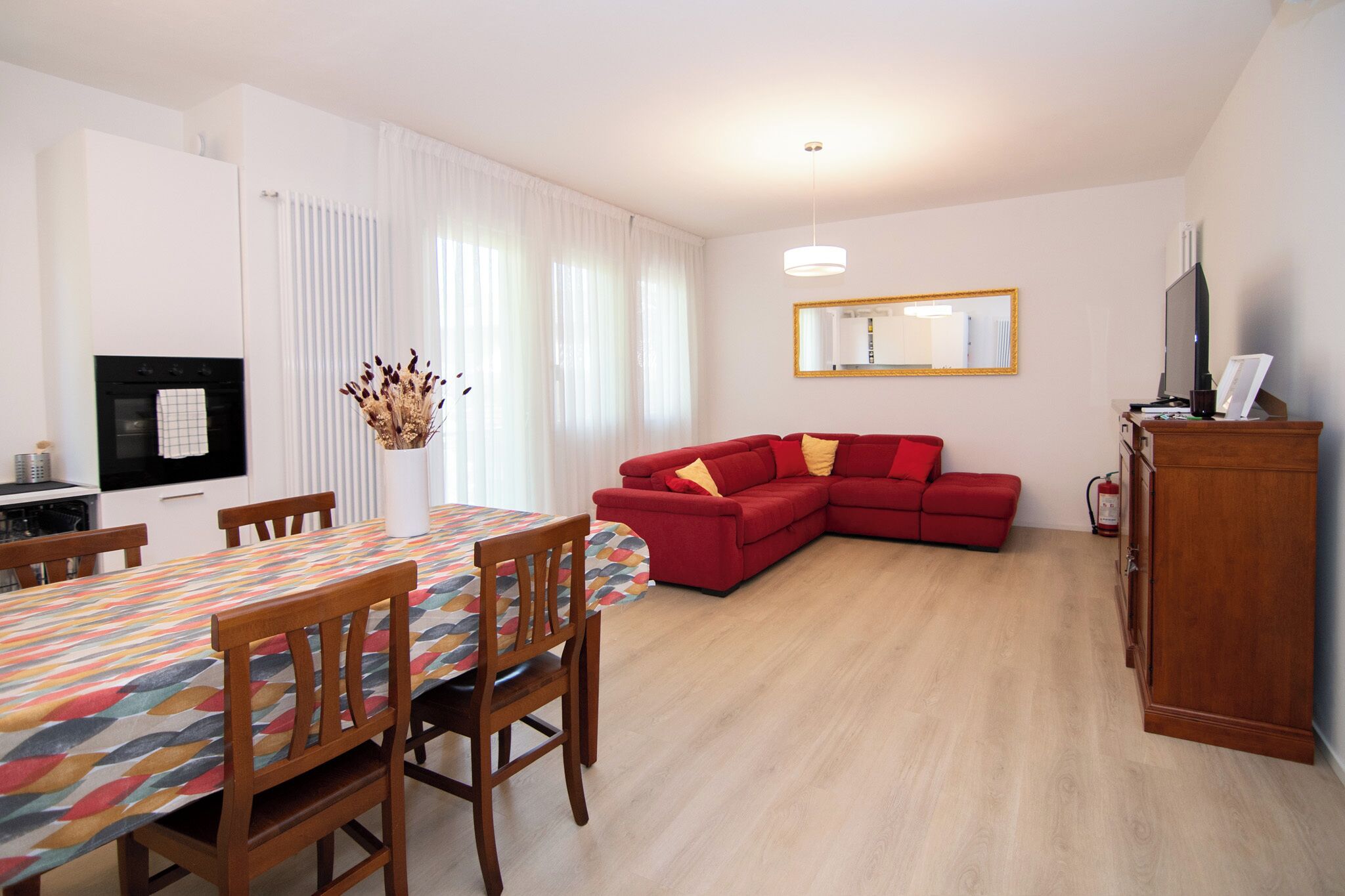 A modern living room with a red sofa, a dining table with chairs, a kitchen area with a stove, and a large mirror on the wall.
