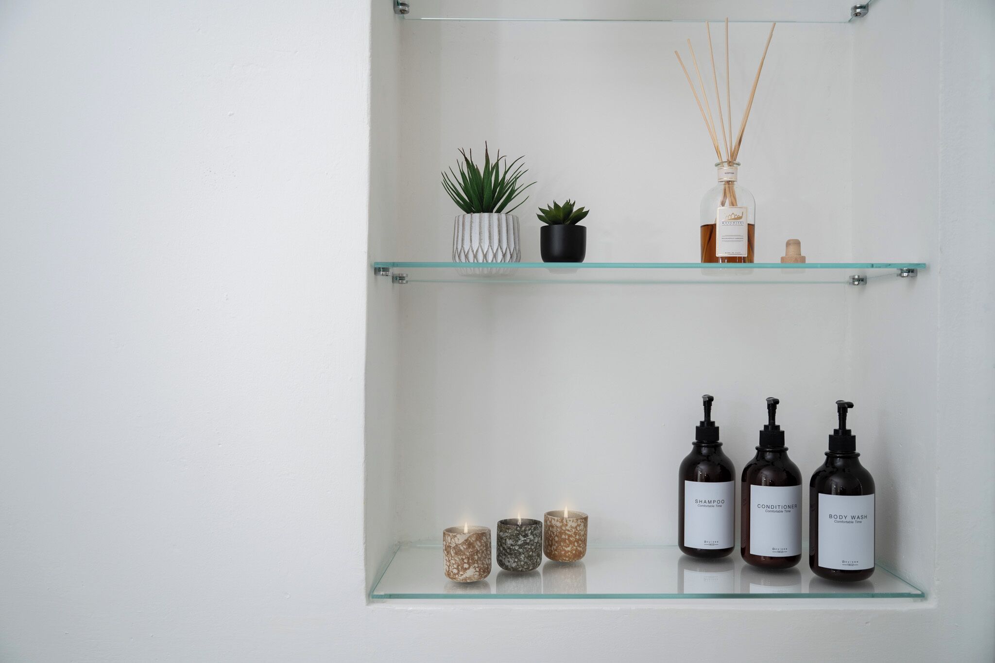 Un baño con dos estantes de vidrio, uno con una planta en maceta, un difusor de cañas y tres botellas de champú, acondicionador y gel de baño.