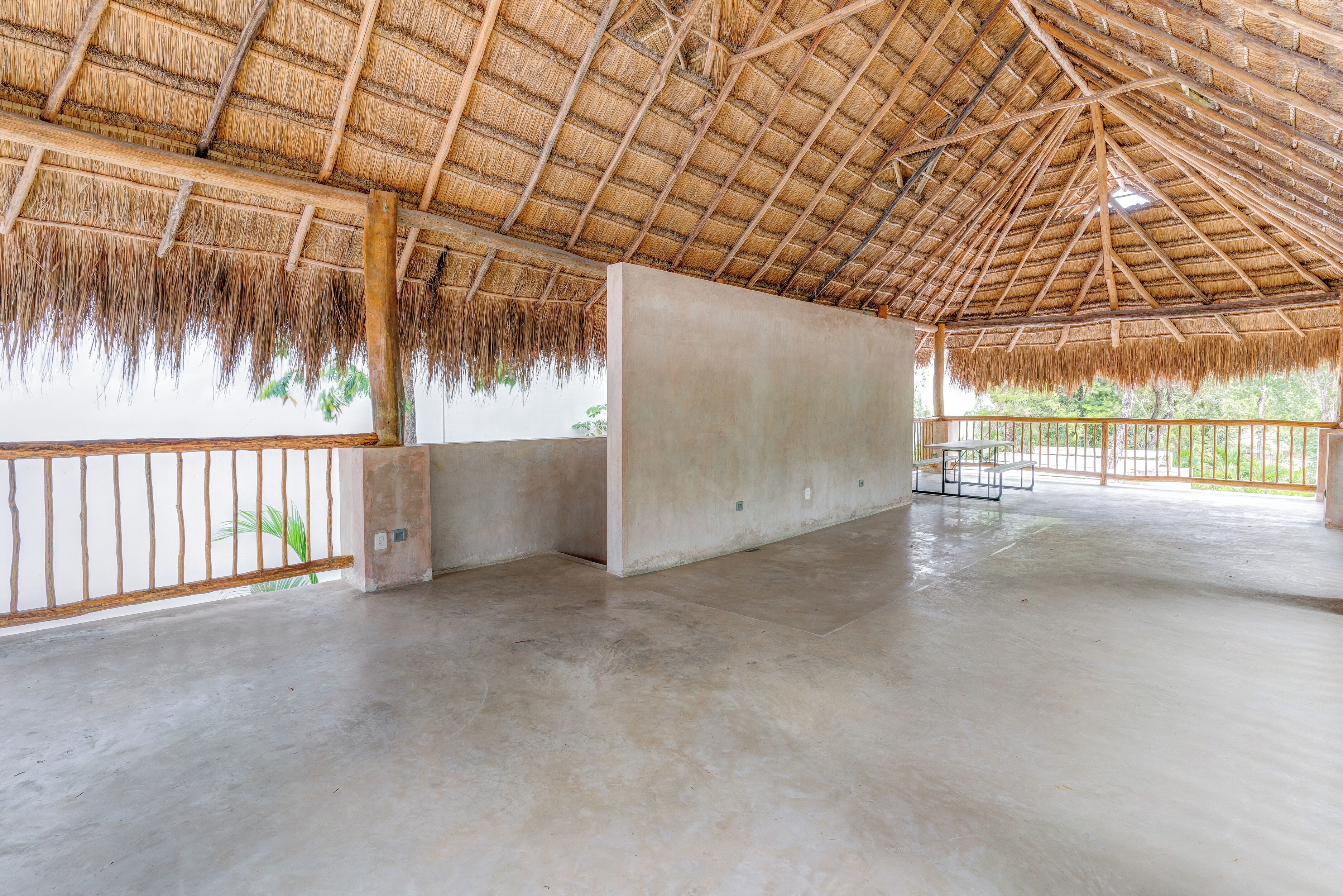 An open, spacious room with a thatched roof and concrete walls, featuring a balcony with a metal railing.