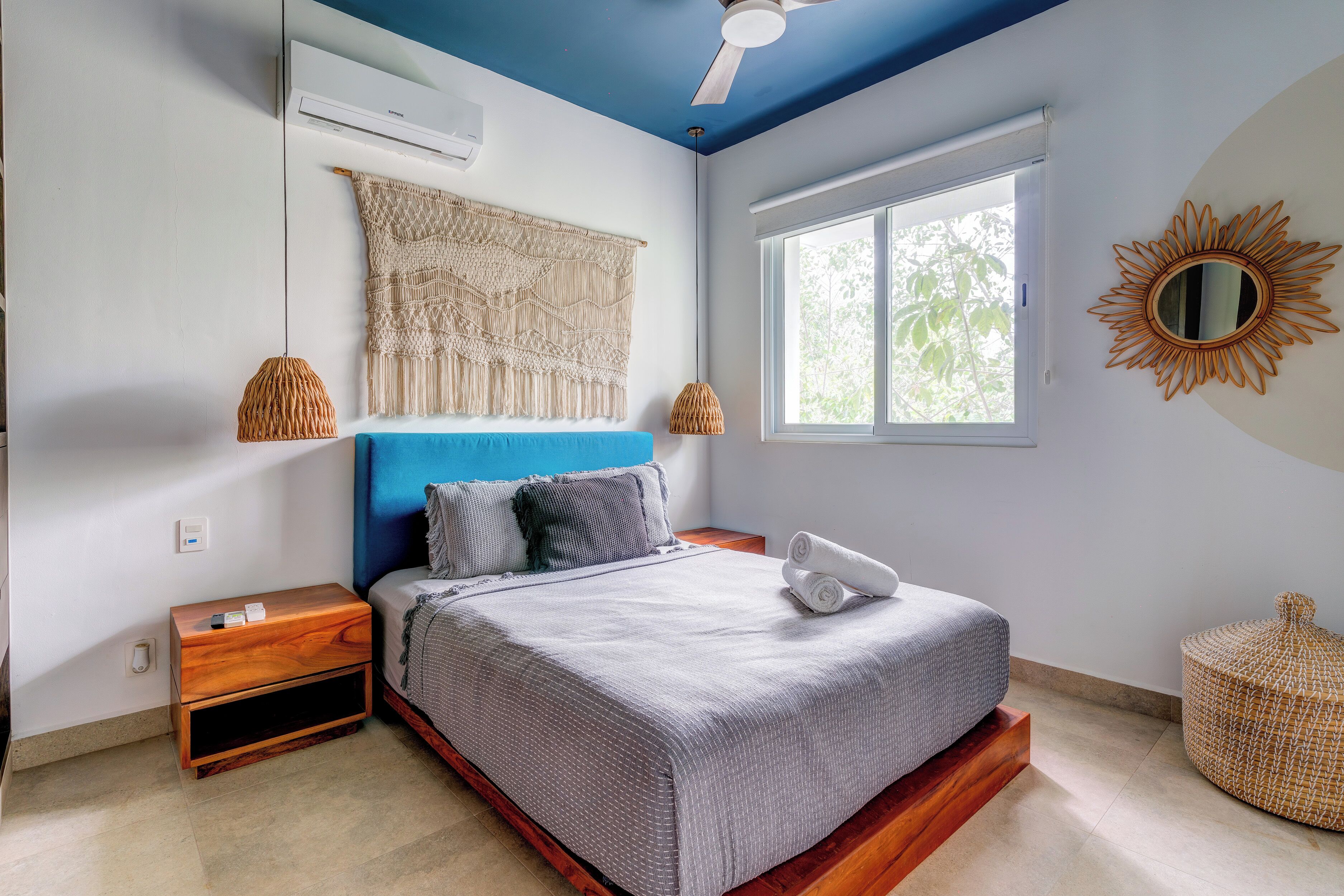 A bedroom with a bed, nightstand, wall-mounted air conditioner, and a window with a view of trees.
