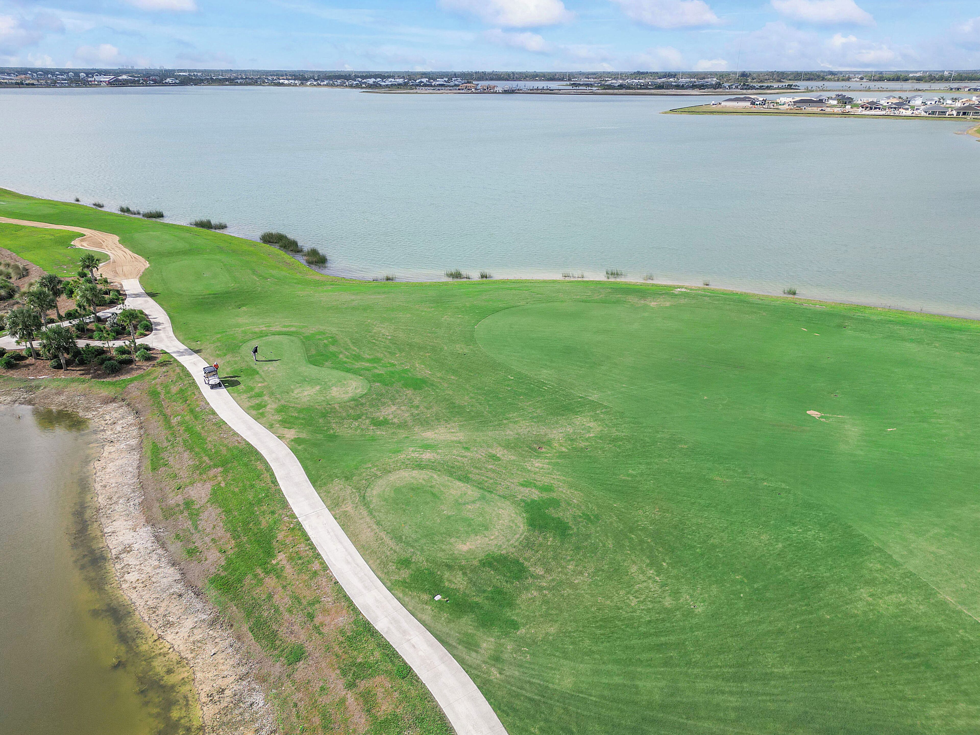 Un campo da golf con specchio d'acqua, vialetto in cemento e una costa lontana con edifici.