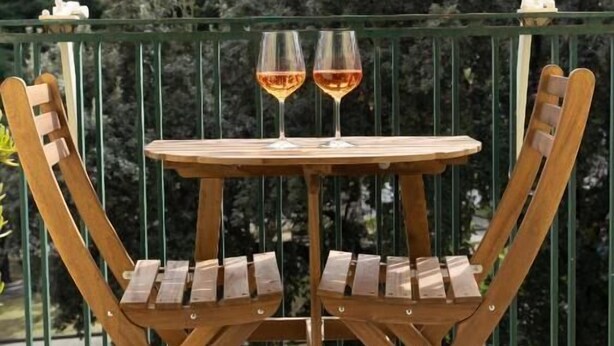 A wooden table with two chairs on a balcony, two glasses of wine, and a railing with greenery in the background. - Sofia e la città delle meraviglie (Naples)