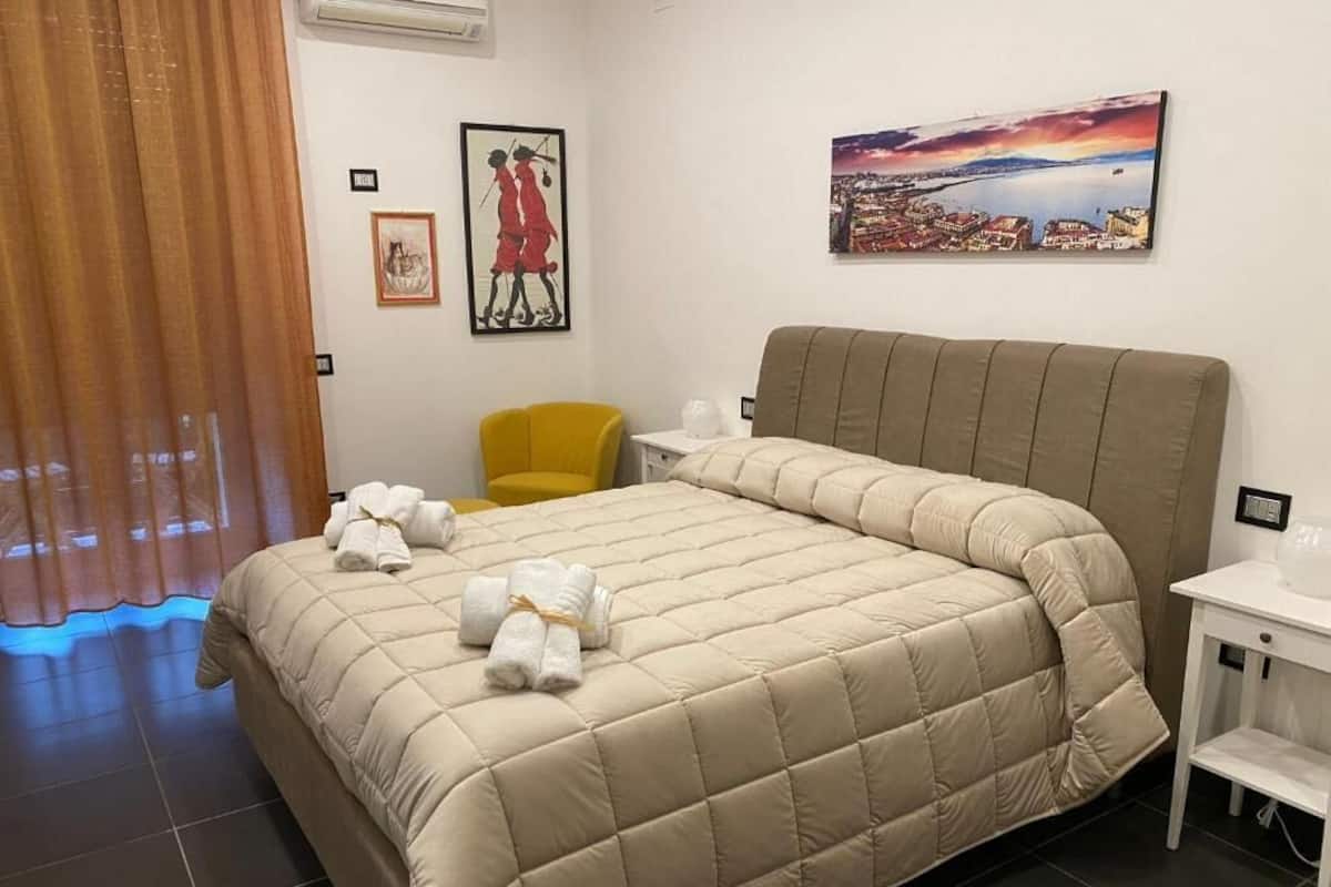 Une chambre d’hôtel avec un grand lit, une petite chaise jaune, une table de chevet et deux tableaux accrochés au mur.