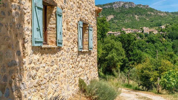 A stone building with blue shutters, a dirt path, and a village in the background. - Le frêne – La Ferme des Aygues, Drôme Provençale (Montaulieu)