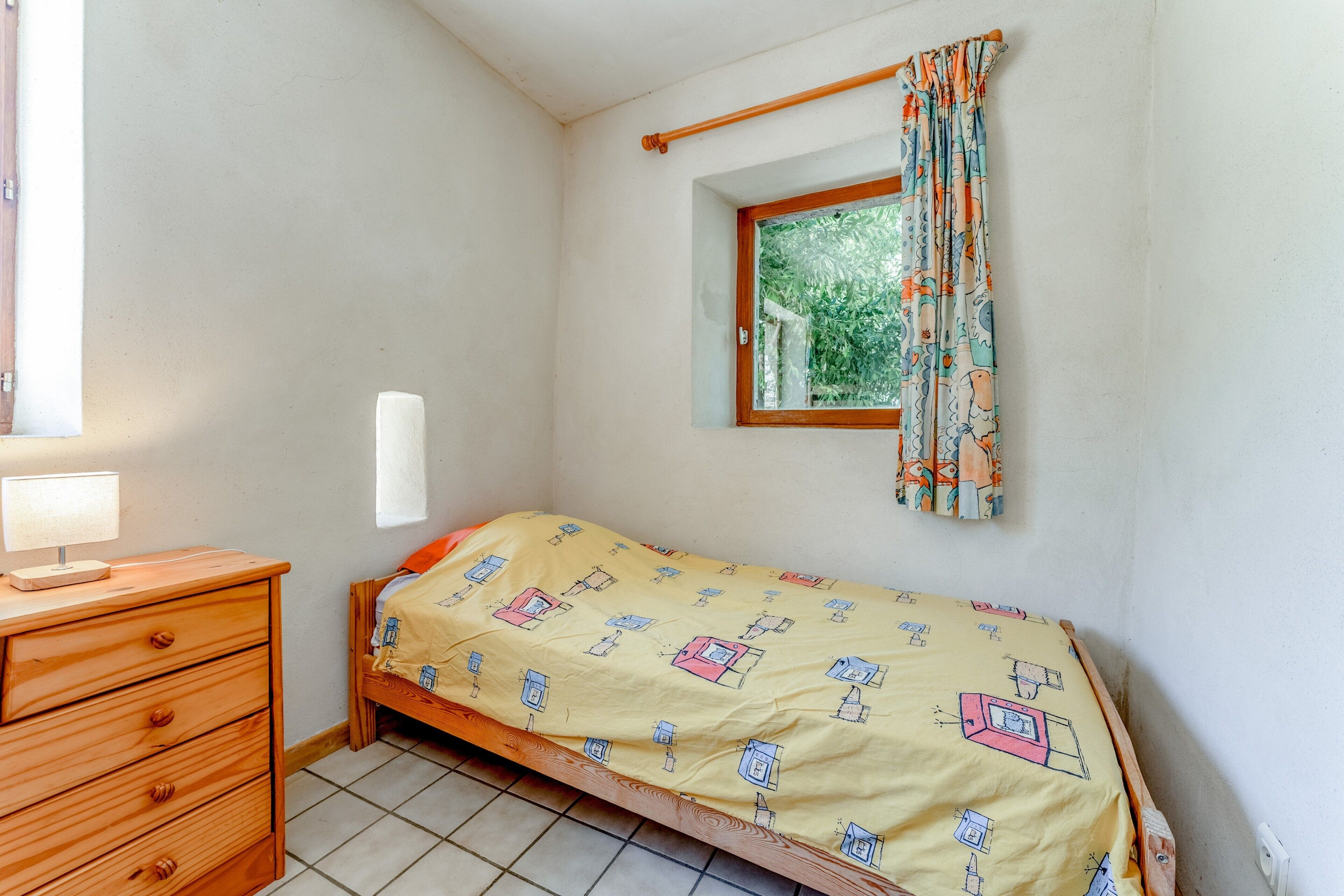 Een kleine slaapkamer met een enkel bed, een houten dressoir, een raam met gordijnen, en een klein raam boven het bed.