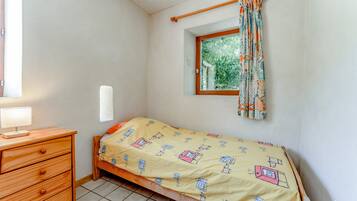 Een kleine slaapkamer met een enkel bed, een houten dressoir, een raam met gordijnen, en een klein raam boven het bed.
