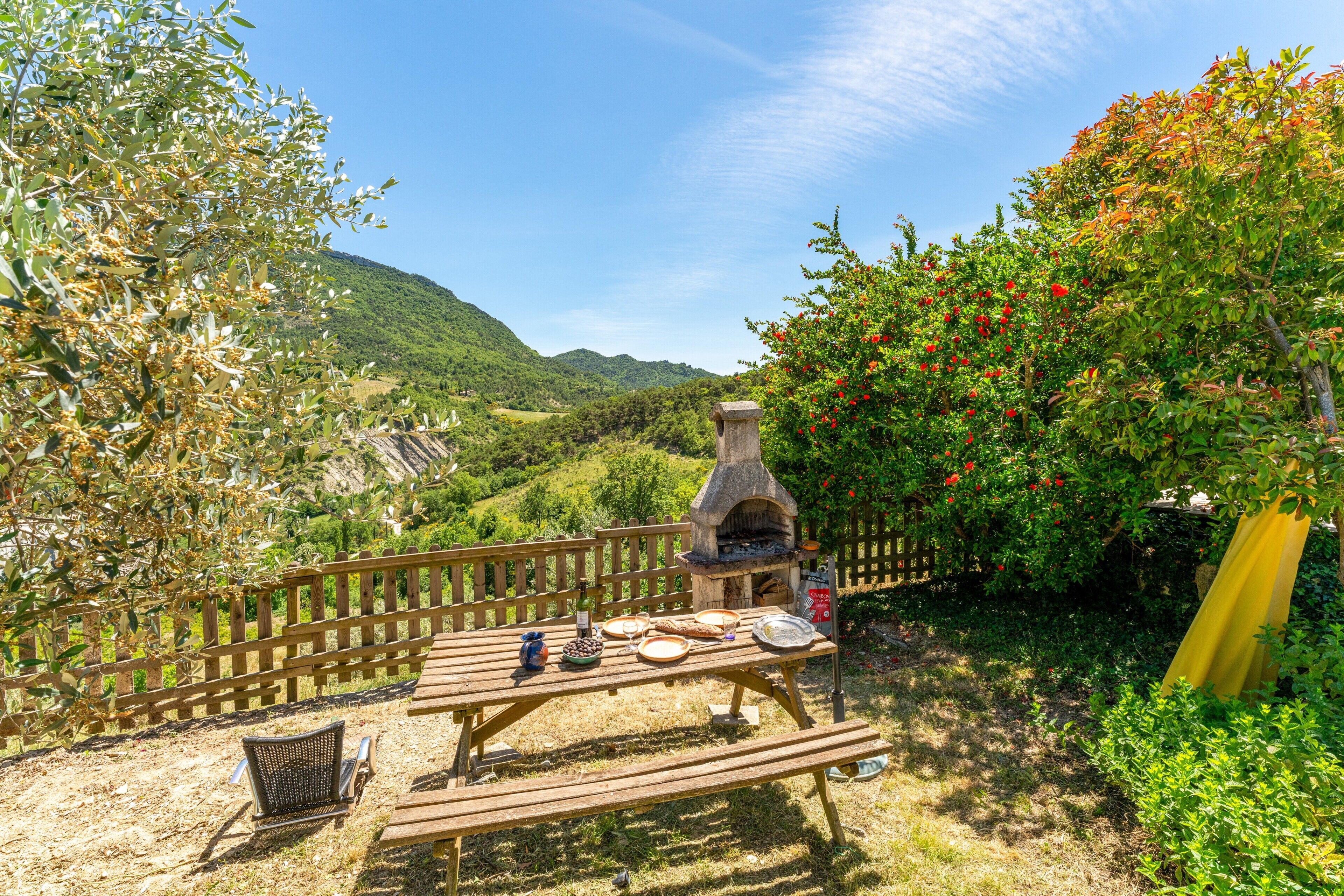Un espace de restauration en plein air doté d’un four en pierre, d’une table et de chaises en bois, le tout entouré de verdure et délimité par une clôture en bois.