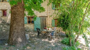 A rustic stone building with a tree in the foreground, outdoor seating, and a small table with items. - Le frêne – La Ferme des Aygues, Drôme Provençale (Montaulieu)