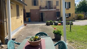 A covered patio area with a table and chairs, potted plants, and a building with a yellow facade and shutters. - Casa Roda – Your Home by Lake Trasimeno, Between Umbria and Tuscany (Castiglione del Lago)