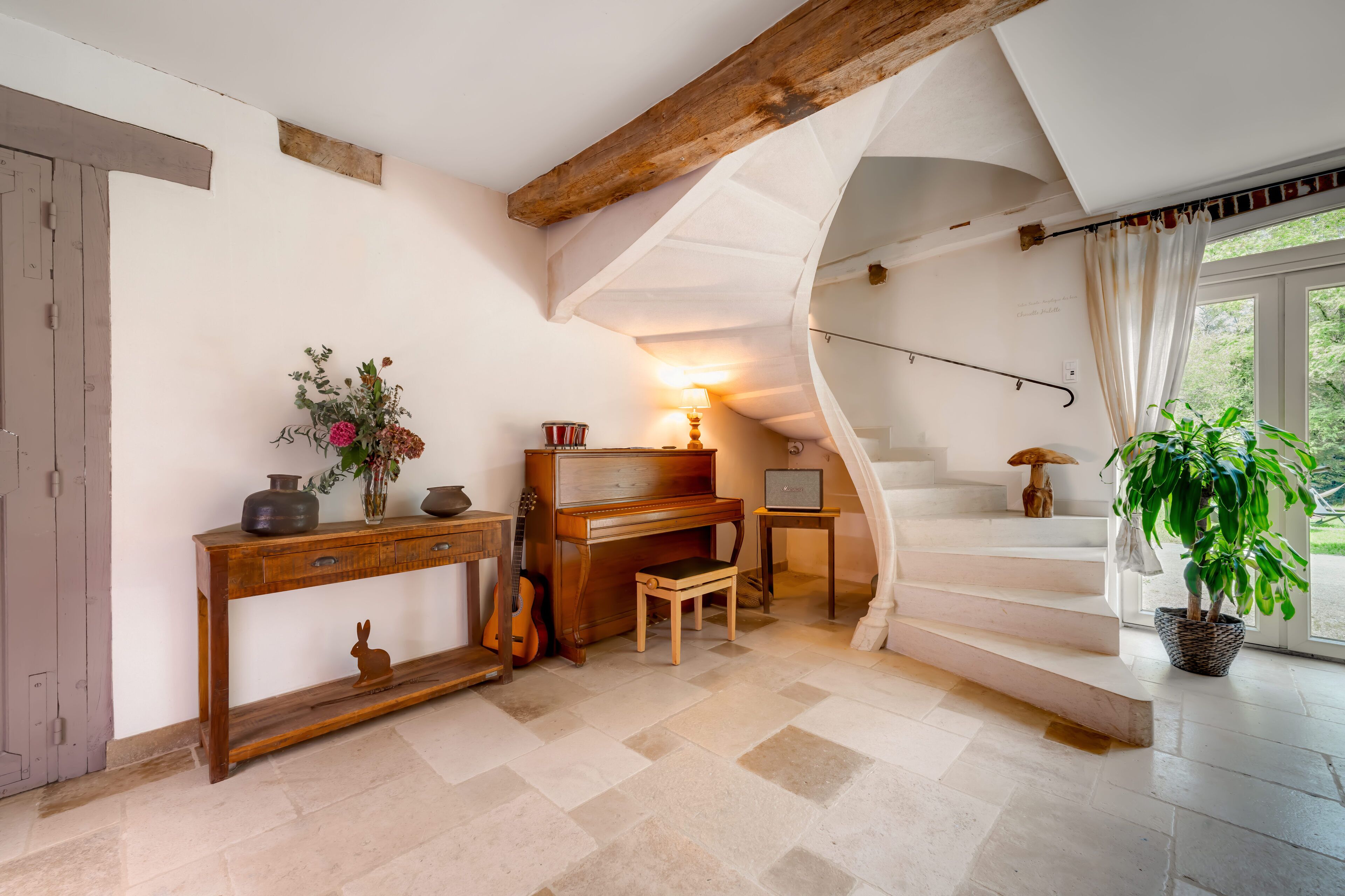 Une pièce chaleureuse avec un piano en bois, une petite table ornée d’un vase de fleurs, une guitare et un escalier menant à un balcon d’où l’on peut admirer la verdure.