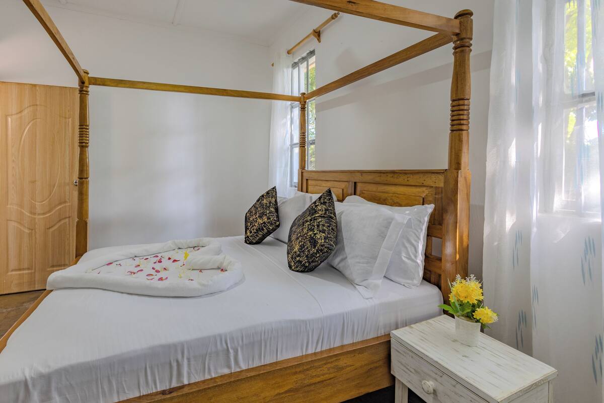 Una cama con dosel, ropa de cama blanca y cojines decorativos, una mesita de noche con un florero y un armario de madera.