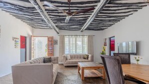 A living room with a stone floor, two sofas, a wooden coffee table, and a dining table with wicker chairs. - AUREGI VILLAS (Diani Beach)