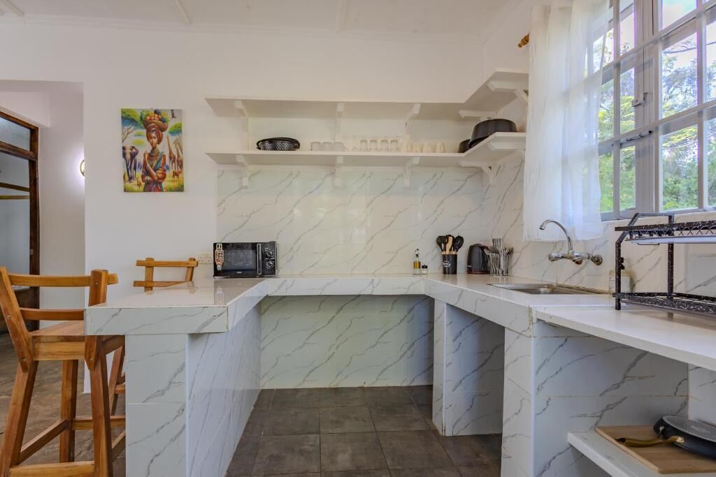 Una cocina con encimera de mármol, microondas y fregadero. Hay una ventana con cortinas y un cuadro en la pared.