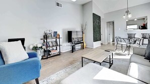 A modern living room with a blue sofa, a coffee table, and a TV. There is a bookshelf with books and decorative items, and a dining area with a table and chairs. - South Austin Retreat: Pet-Friendly Townhouse (Austin)