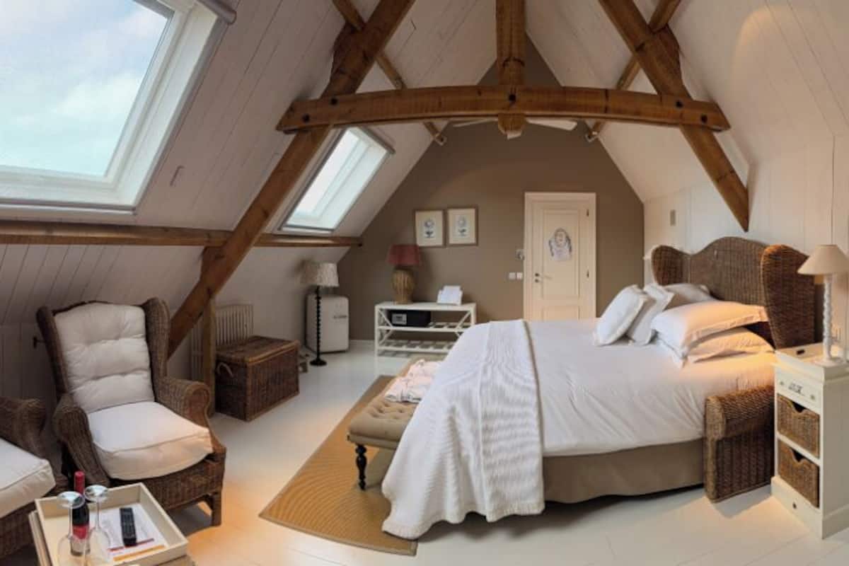 Een gezellig slaapkamer onder het dak, met een schuine kap, een bed met witte lakens, twee gestoffeerde fauteuils, een nachtkastje en een kleine bedlamp.