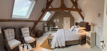 A cozy attic bedroom with a sloped ceiling, a bed with white linens, two upholstered chairs, a bedside table, and a small bedside lamp.