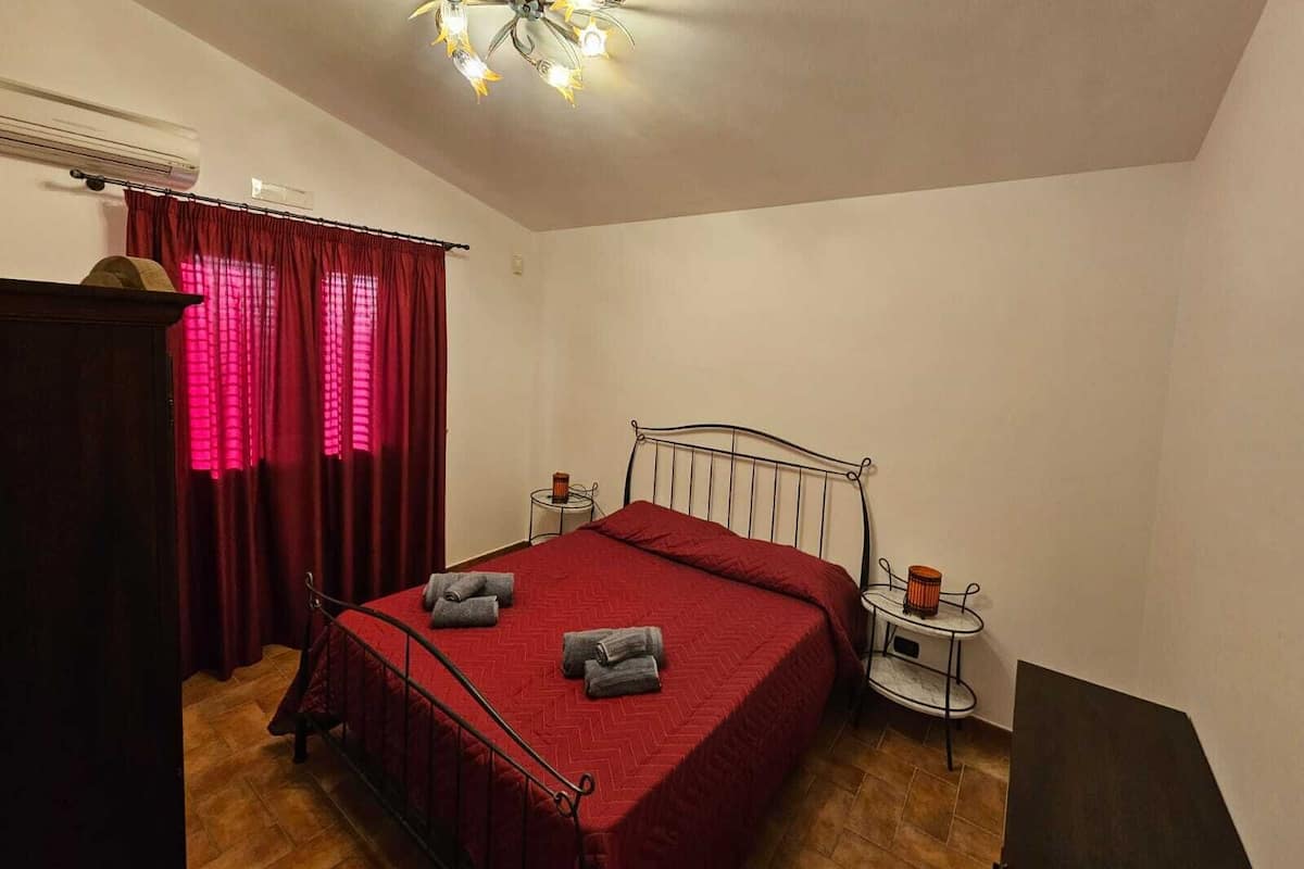 A bedroom with a metal bed frame, a red bedspread, a chandelier, a small bedside table with a lamp, and a wardrobe.