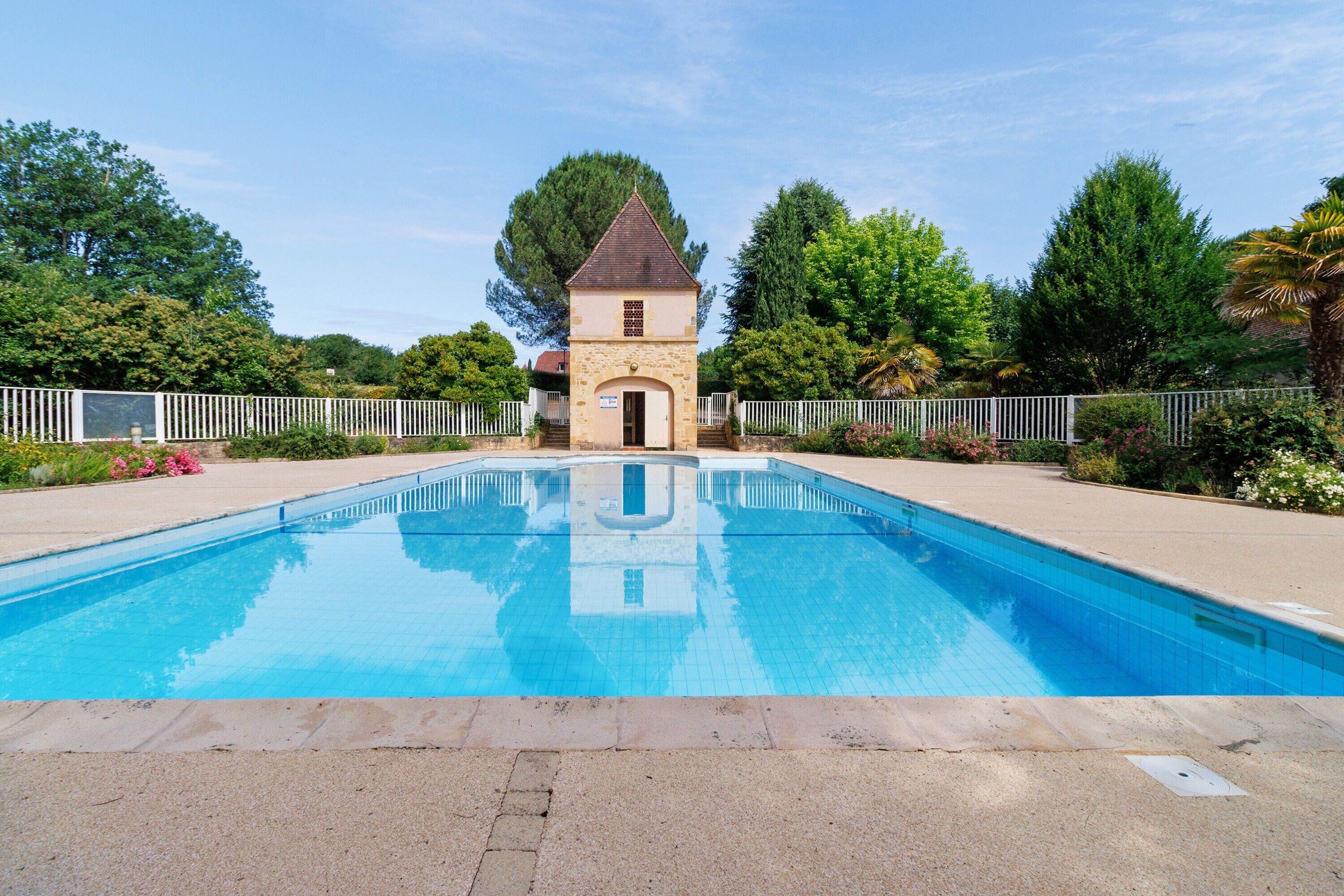 Une piscine avec un petit bâtiment en pierre au centre, entourée d’arbres et d’une clôture blanche.