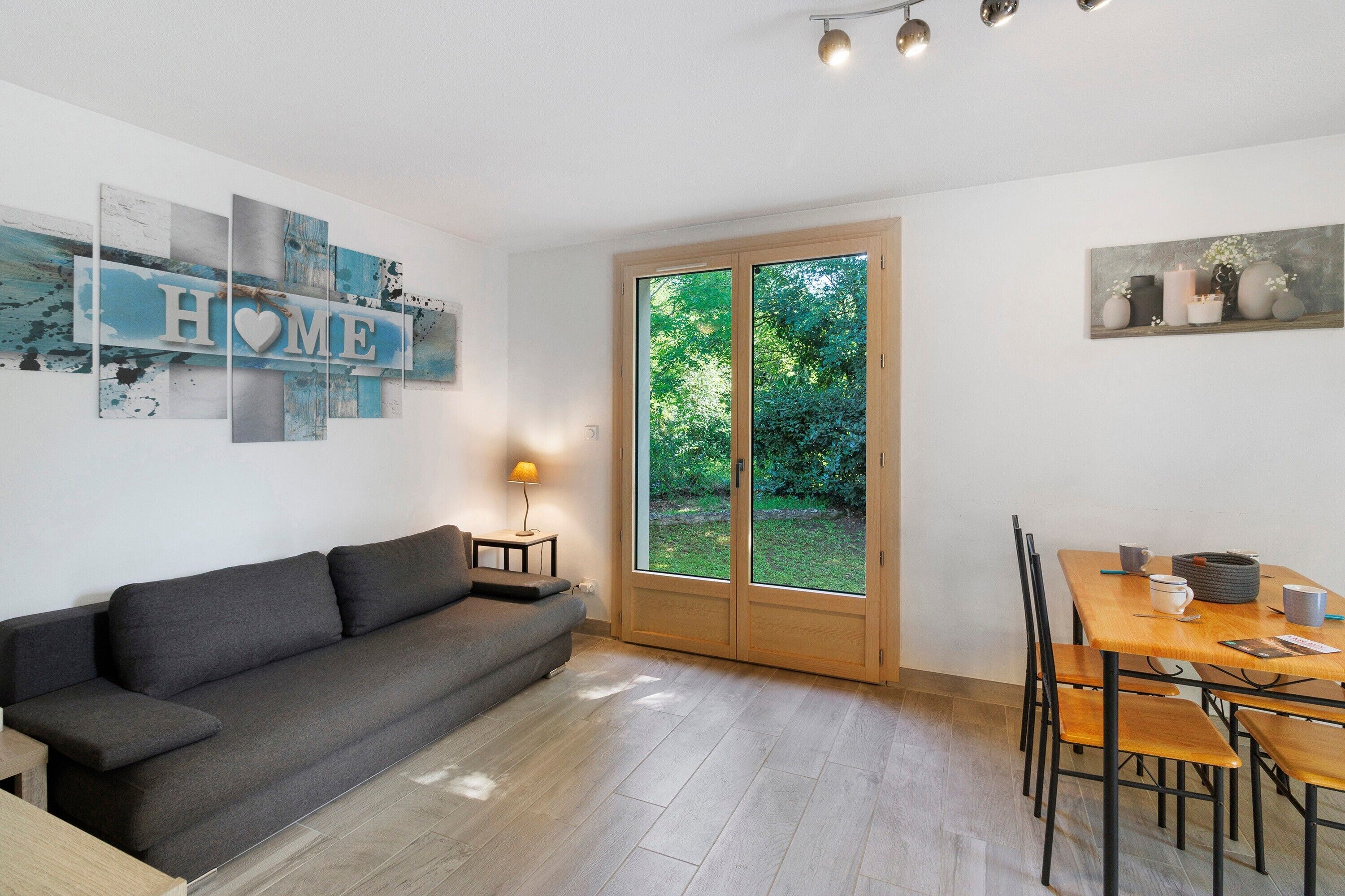 A modern living room with a grey sofa, a dining table with chairs, a lamp, and a large window with a view of greenery.