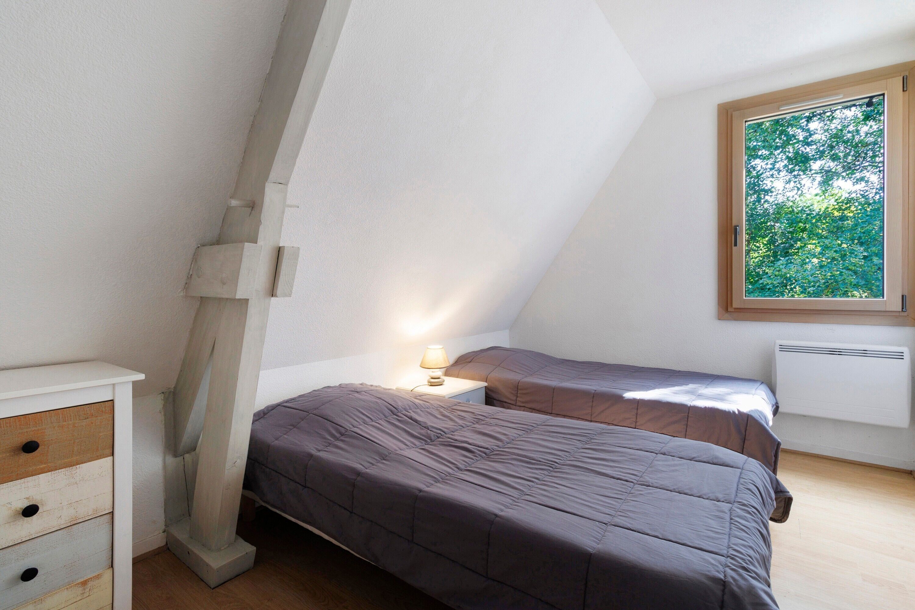 Een slaapkamer met twee bedden, een nachtkastje, een dressoir en een raam met uitzicht op bomen.
