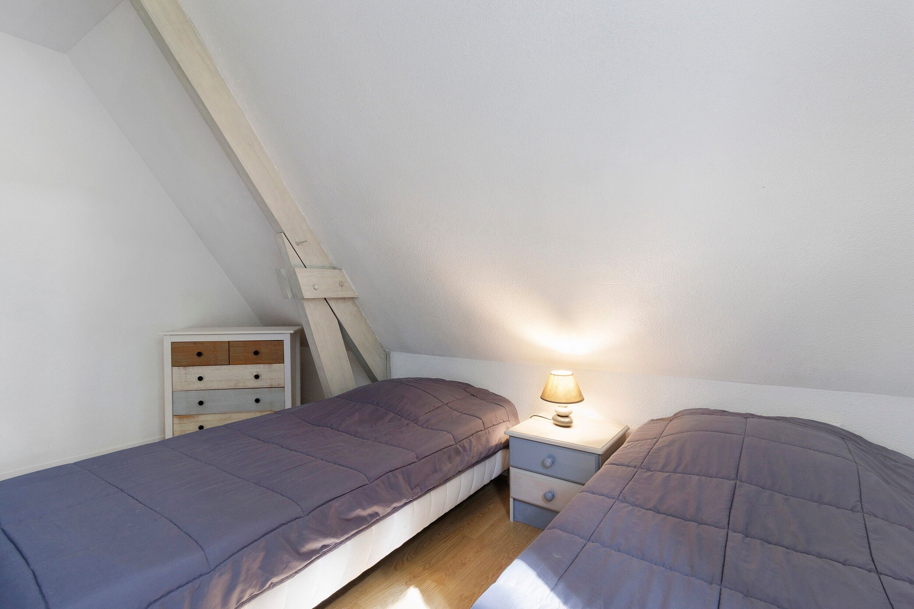 Una habitación pequeña con techo inclinado, dos camas, una mesita de noche con lámpara y un aparador de madera.