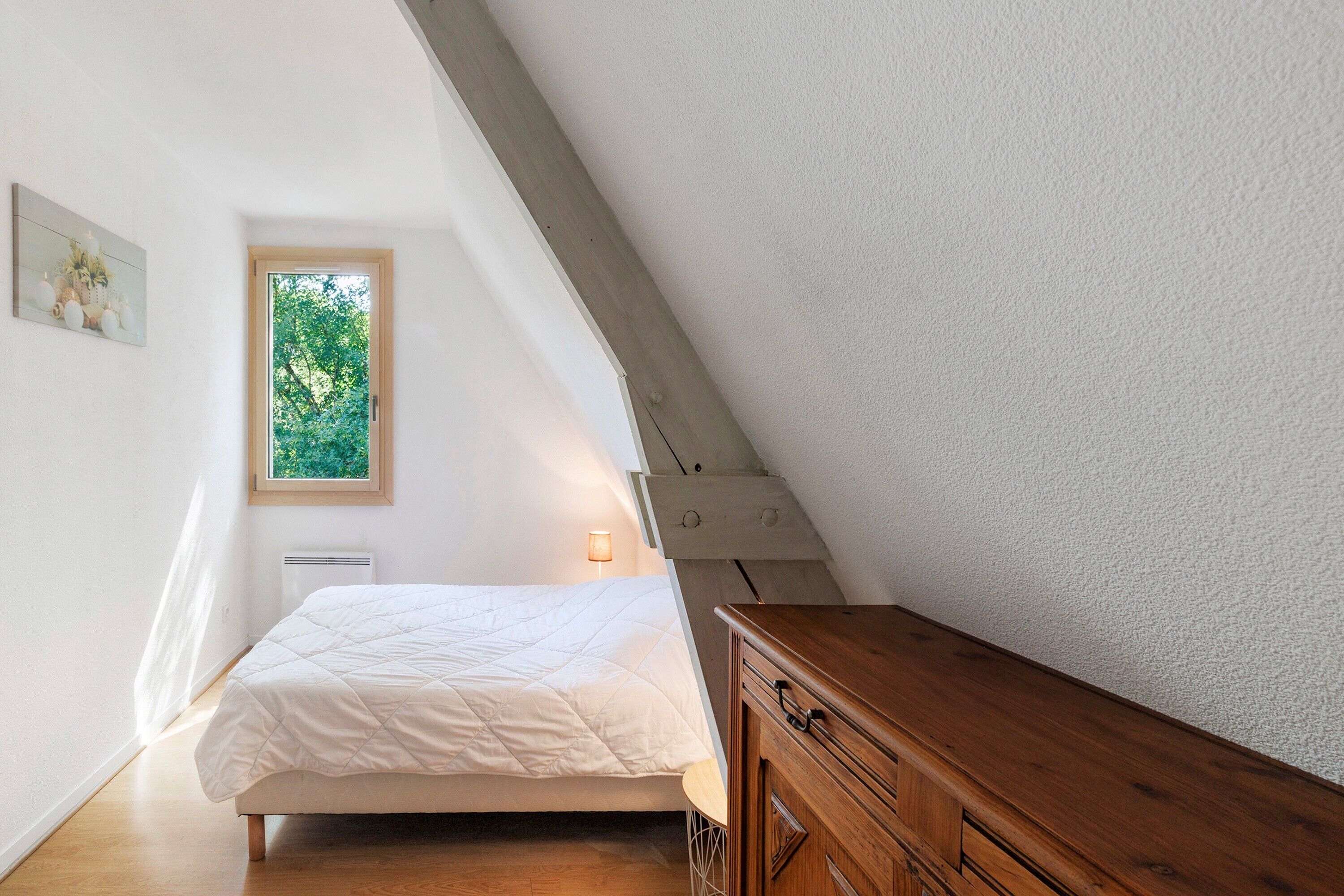 Une chambre à coucher avec un lit, une commode en bois, une fenêtre donnant sur un paysage verdoyant et une lampe fixée au mur.