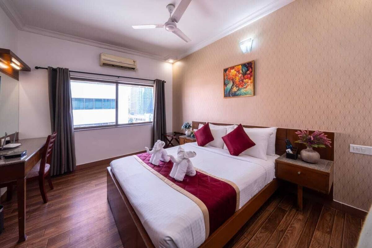 A hotel room with a large bed, a desk, and a chair. There is a window with curtains, a ceiling fan, and a painting on the wall.