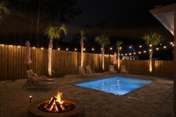 A backyard with a swimming pool, lounge chairs, a fire pit, and string lights.