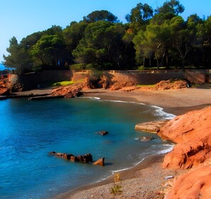 A coastal beach with clear blue water, rocky shoreline, and dense green trees in the background. - Sunny Break – Cozy Retreat Steps from the Sea (Saint-Raphaël)
