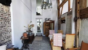 Un interior rústico con vigas de madera a la vista, pared de ladrillo, un banco de madera, una silla de mimbre, un armario de madera y una lámpara de metal colgante.