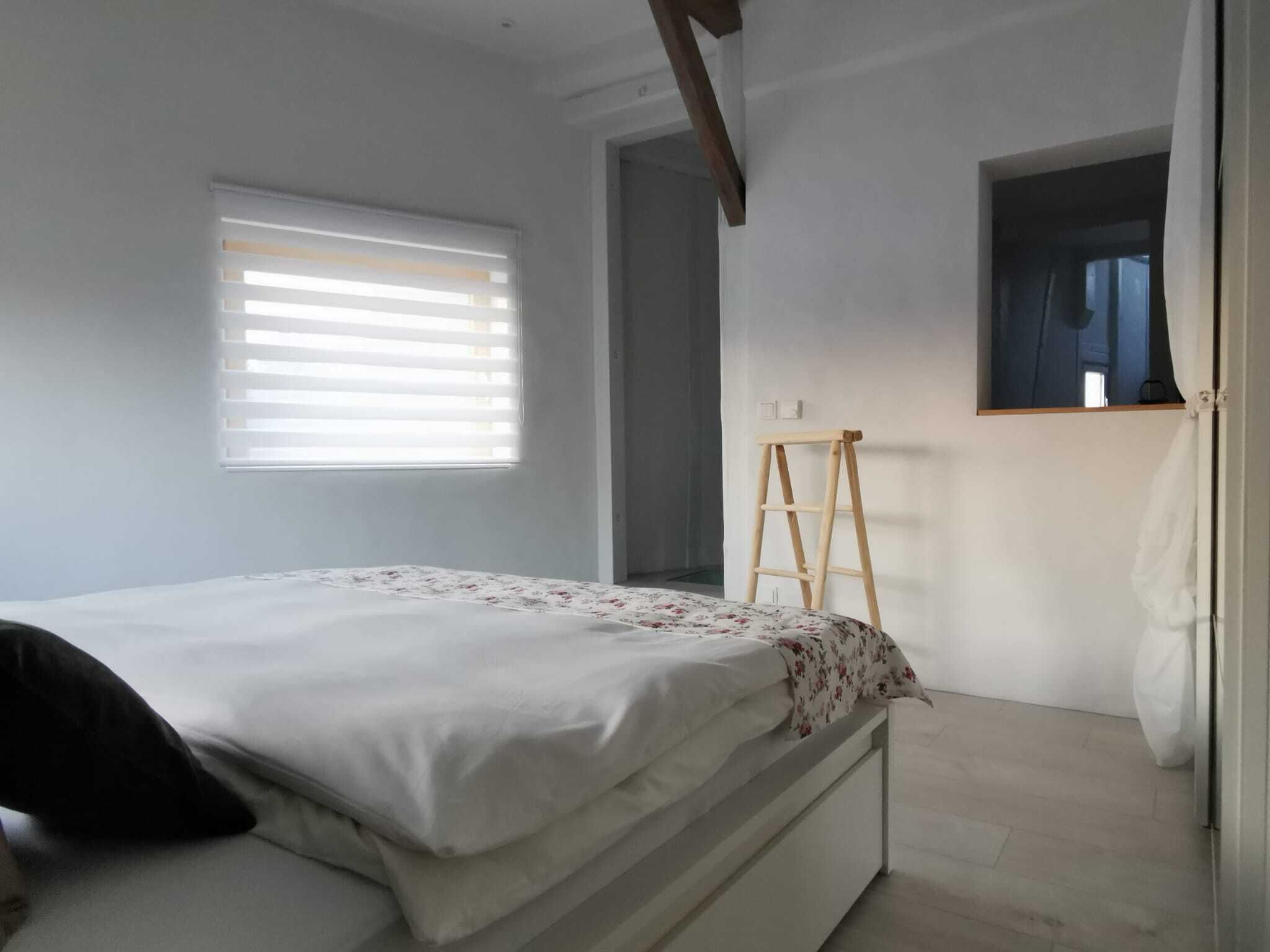 Un dormitorio con cama, ventana con persianas, escalera y puerta que da a otra habitación.
