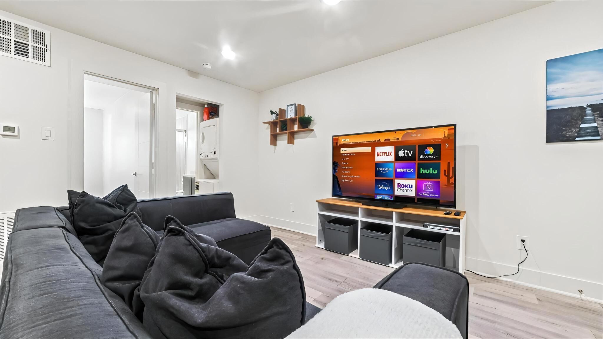 Un salon moderne doté d’un téléviseur à écran plat diffusant des services de streaming, d’un canapé gris avec des coussins noirs et d’un meuble de rangement en bois.