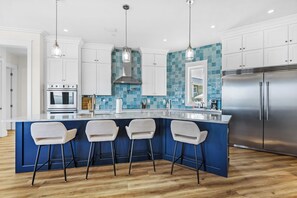 A modern kitchen with a central island, bar stools, stainless steel appliances, and a blue-tiled backsplash. - 🌞🌴Sound Views | Golf Cart | Community Pool | An Island in the Sun🌞🌴 (Navarre)