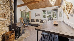 A rustic interior with a stone fireplace, wooden beams, a sofa, and a dining table. - Ptarmigan Condo w/ Loft and Fireplace (Whitefish)