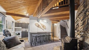 A cozy living area with a sofa, a fan, a fireplace, and a kitchen in the background. - Ptarmigan Condo w/ Loft and Fireplace (Whitefish)