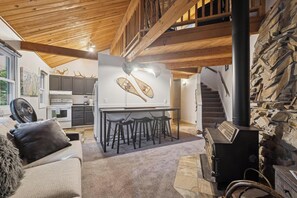 A cozy living area with a sofa, a fan, a fireplace, and a kitchen in the background. - Ptarmigan Condo w/ Loft and Fireplace (Whitefish)