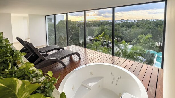 A modern balcony with a hot tub, lounge chairs, and a view of a cityscape and greenery. - Luxury Condo in PGA Golf Resort | Bahia Principe (Akumal)